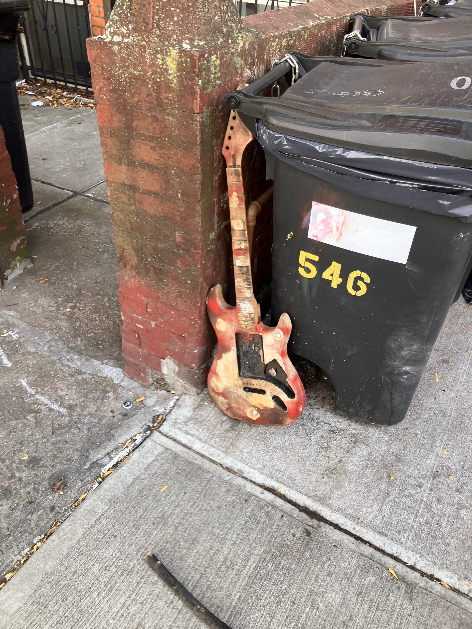 A worn guitar leaning beside a rubbish bin on the sidewalk